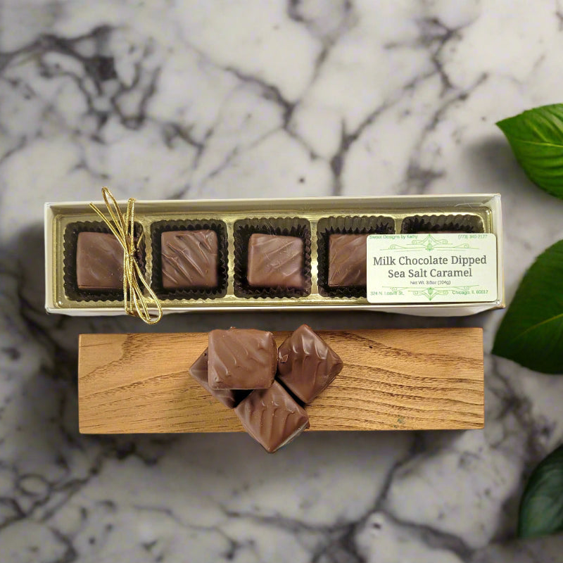 A box of chocolate dipped sea salt caramels placed on a wooden board with three additional caramels outside the box, tied with a string.