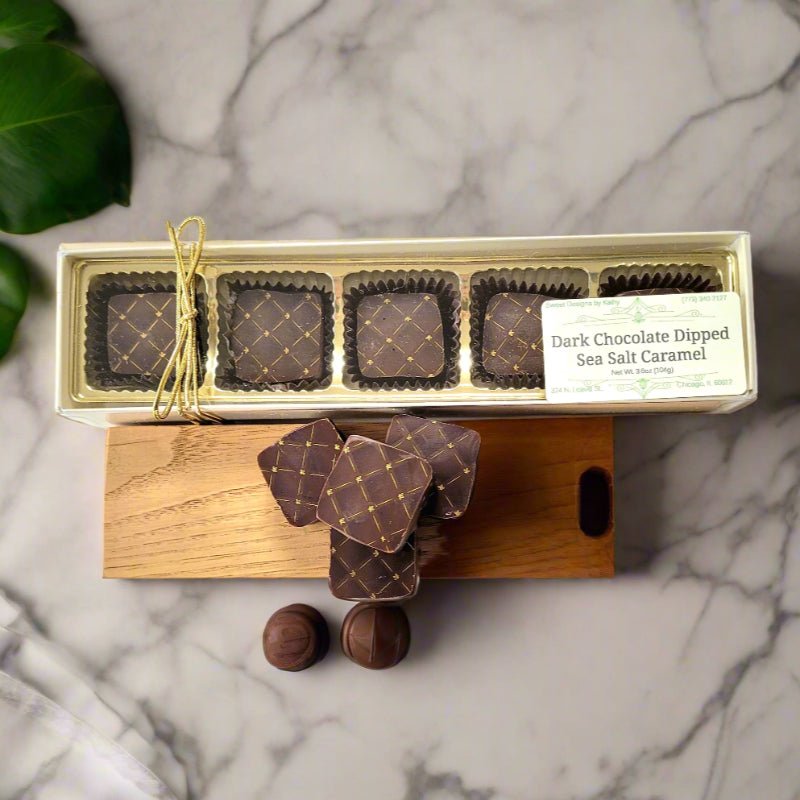 A box of chocolate dipped sea salt caramels placed on a wooden board with three additional caramels outside the box, tied with a string.